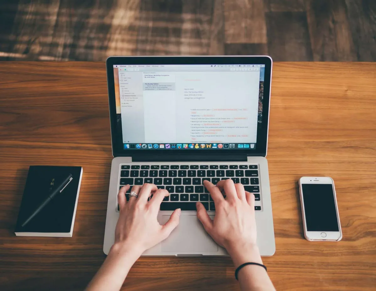 Overhead view of a person typing on a laptop at a wooden desk beside a notebook, pen and phone